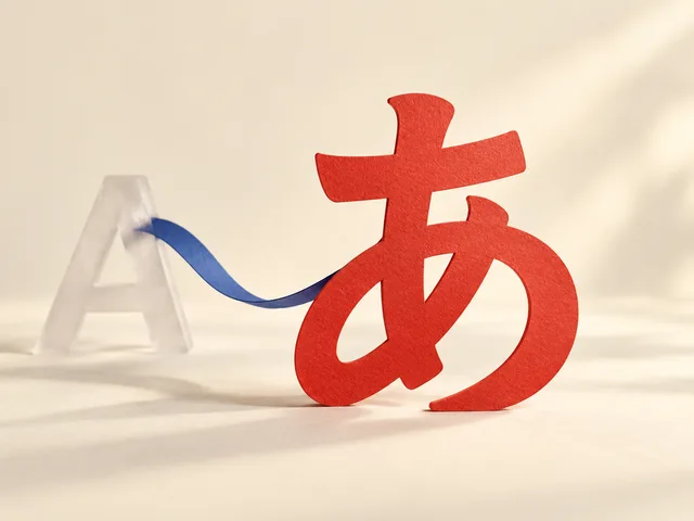 Sculpture de papier : un hiragana あ vermillon se tient au premier plan, relié par un ruban bleu à une lettre A translucide en retrait.